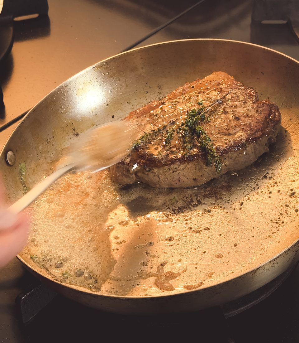 Carne selando na frigideira de inox enquanto é regada com manteiga e tomilho.