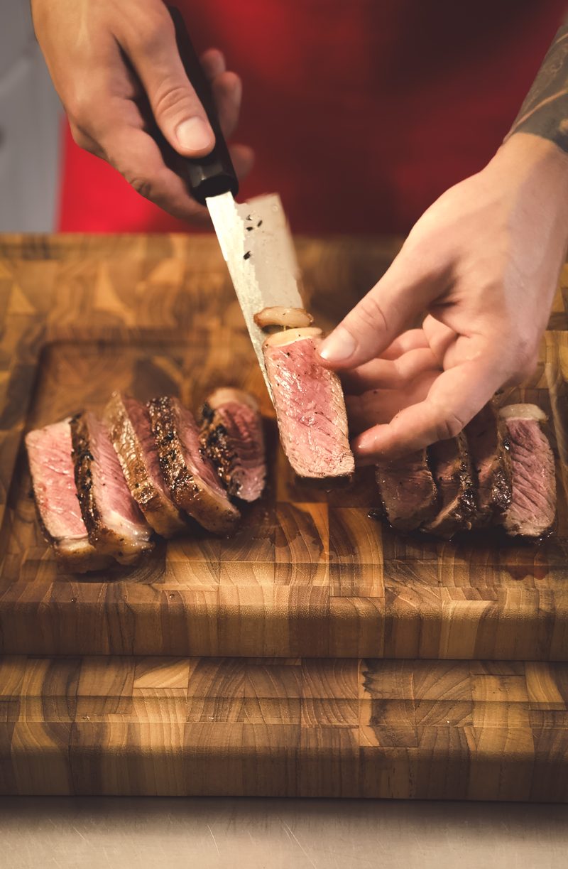 Mãos fatiando uma carne na tábua de madeira, com o ponto rosado à mostra.