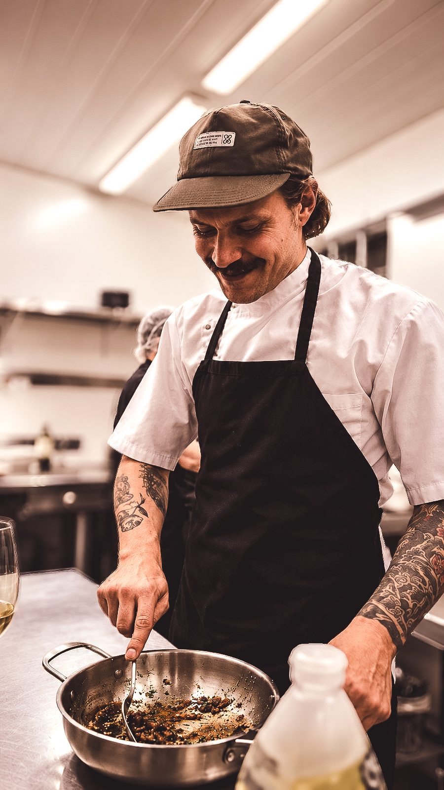 Francisco Dal Medico sorrindo na cozinha, de boné, dólmã branca e avental preto, mexendo uma frigideira.